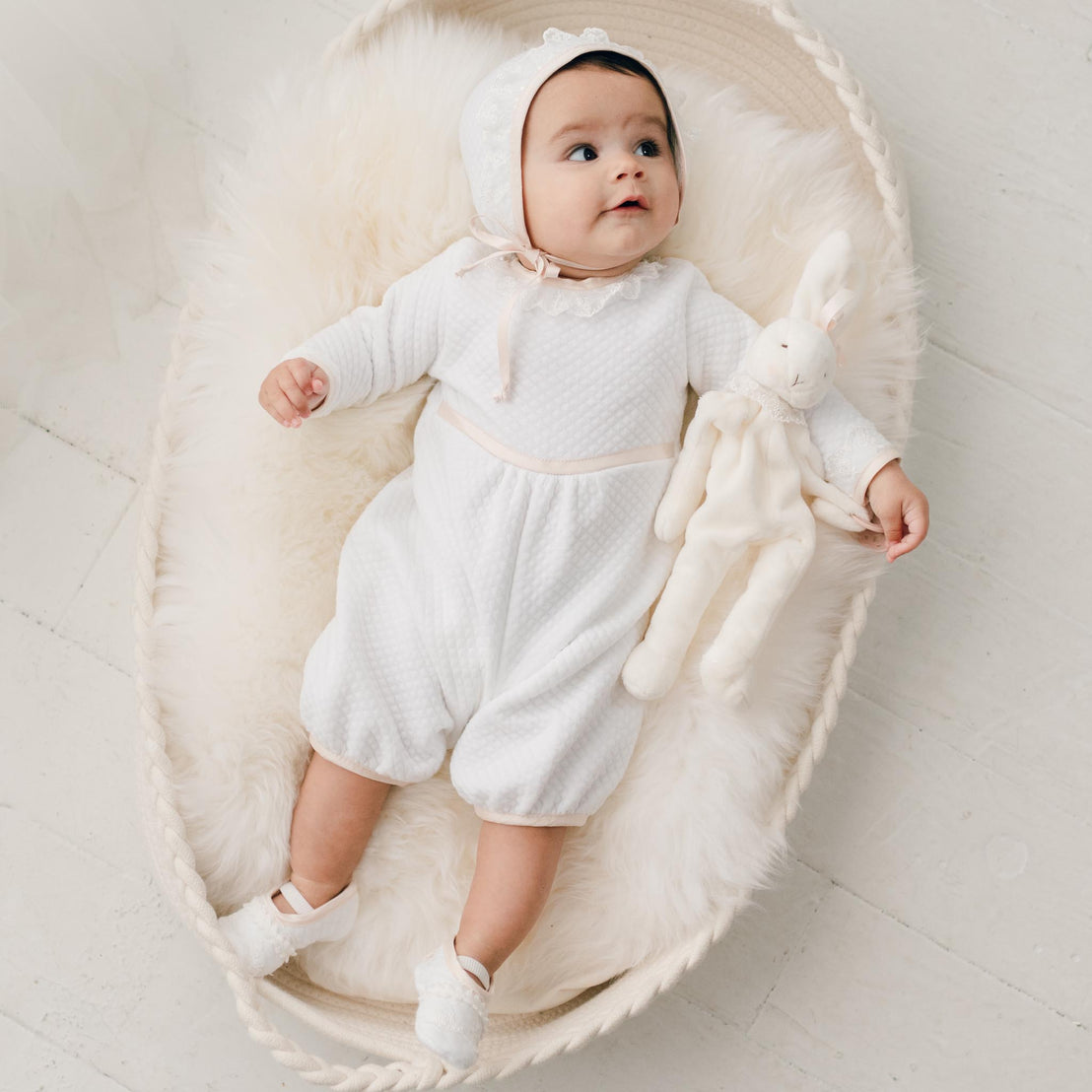 Newborn baby girl sitting in a crib and wearing the Tessa Quilt Romper and Tessa Quilt Bonnet. The romper is made from a white quilted cotton and features ivory Venice Lace along the neck and sleeves and pink champagne silk trim at the cuffs, waist and neck.