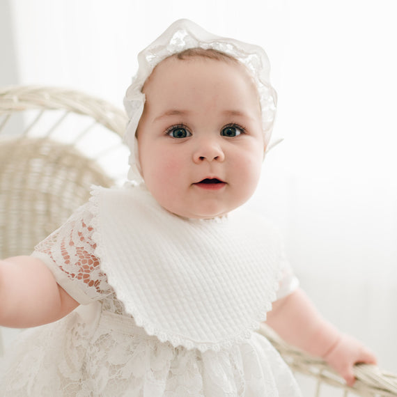 A baby with wide blue eyes and a white headband sits in a rattan chair, wearing an ivory quilted cotton dress and a Rose Christening Bib, looking directly at the camera with a curious expression.
