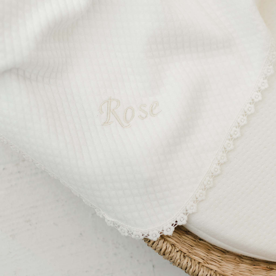 A white, textured Rose Personalized Blanket with the name "Rose" embroidered in elegant cursive, accented with a delicate lace trim, partially resting in a wicker basket on a light surface.