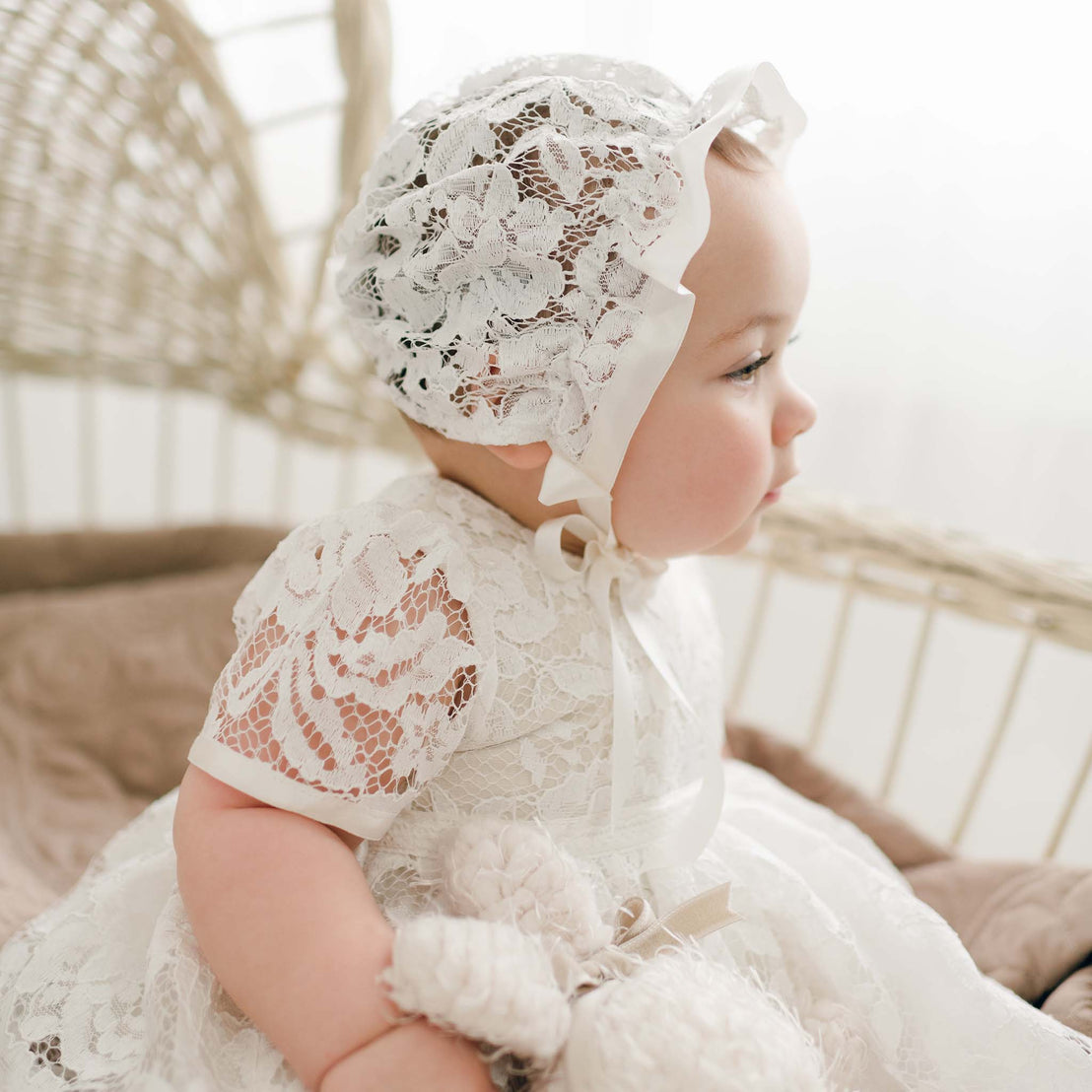 A baby wearing a lace dress and a Rose Lace Bonnet sits in a wicker chair, gazing to the side with a thoughtful expression. The setting is softly lit, creating a warm, gentle atmosphere.