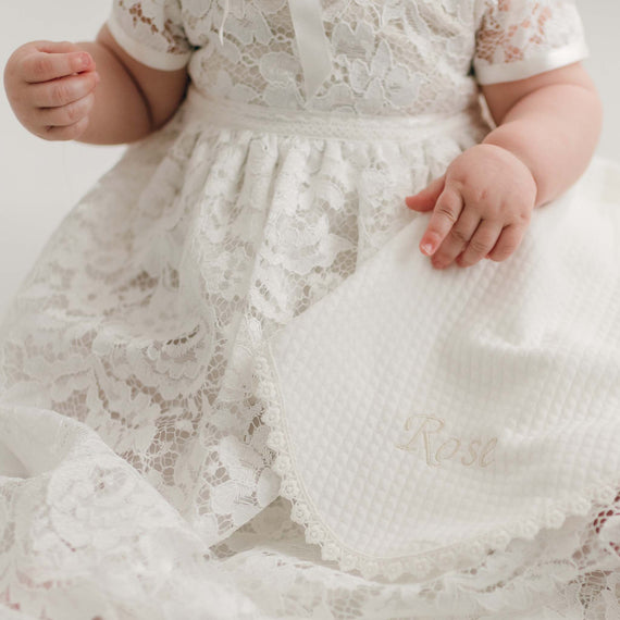 A close-up image of a baby in a white lace dress, holding a Rose Personalized Blanket embroidered with the name "Rose." Only the baby's hands and torso are visible.