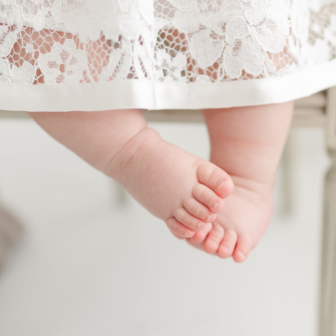 Close-up of a baby's bare feet hanging down, with toes pointing downwards, beneath the hem of the Rose Romper Dress. The background is bright and softly focused.