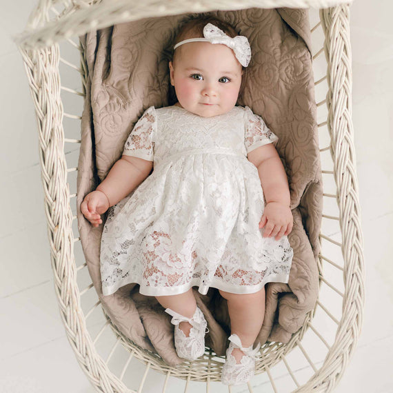 A baby wearing the Rose Romper Dress and matching Rose Bow Headband and Booties lies in an oval, wicker bassinet lined with a beige cushion. The baby looks upward with a curious gaze.