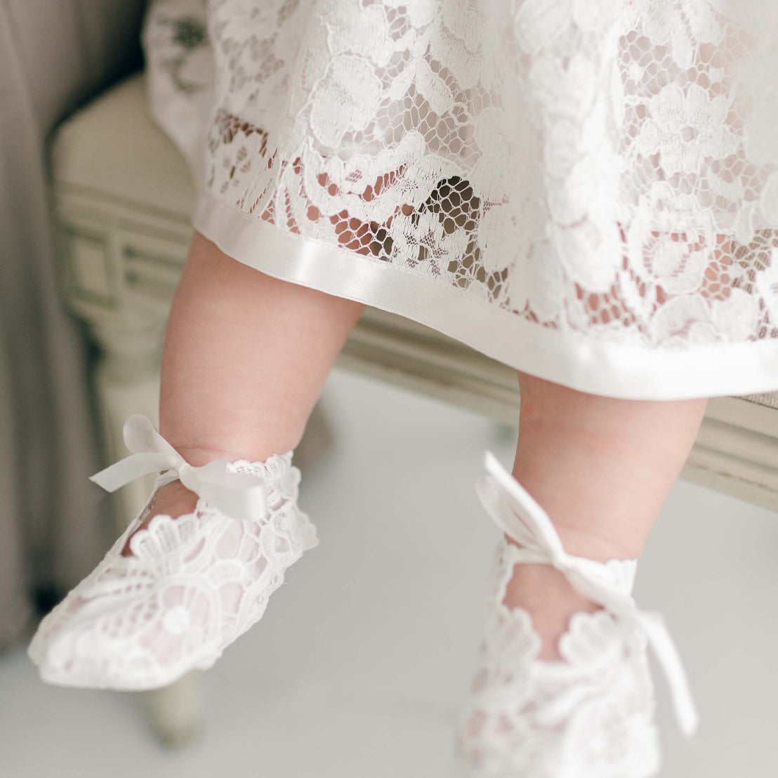 Close-up of a baby's legs wearing white Rose booties with silk bow ties, showcasing the detailed pattern of the lace on a soft-focus background.