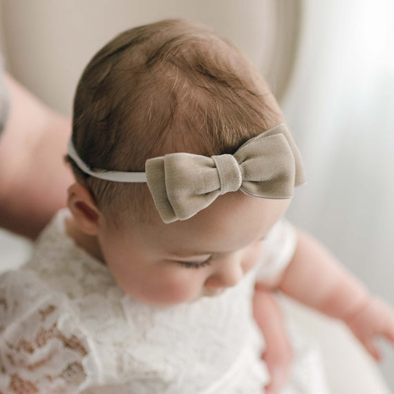 A close-up of an infant wearing a white lace dress and a Rose Velvet Bow Headband, beautifully focusing on the texture of the hair and fabric. The background is softly blurred.