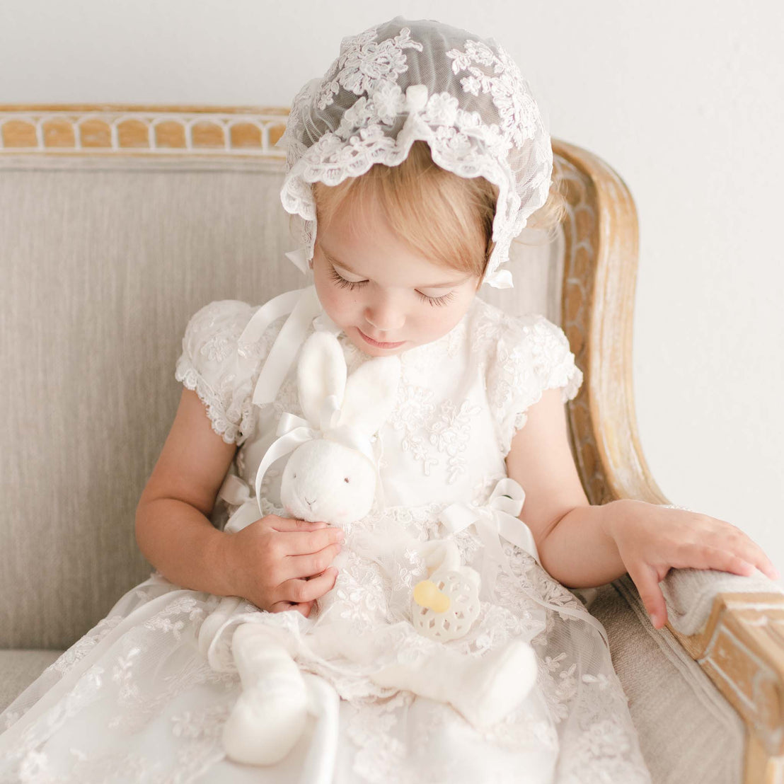 Baby girl lovingly holding a bunny pacifier holder toy. Girl is wearing a christening gown and bonnet.