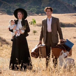 A woman holding a baby and a man with luggage stand in a dry, grassy landscape. The woman wears a long dress and hat, while the man is in a suit. A small dog is beside them. The scene suggests travel or arrival in a rural area.