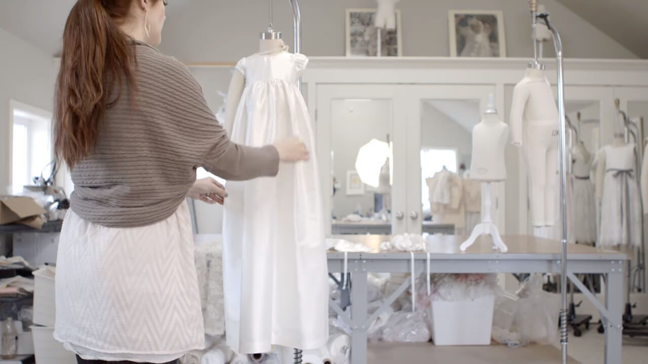 A woman with long hair is adjusting a white dress on a mannequin in a sewing room. The room is filled with sewing supplies, including tables, mannequins, and framed pictures on the wall.