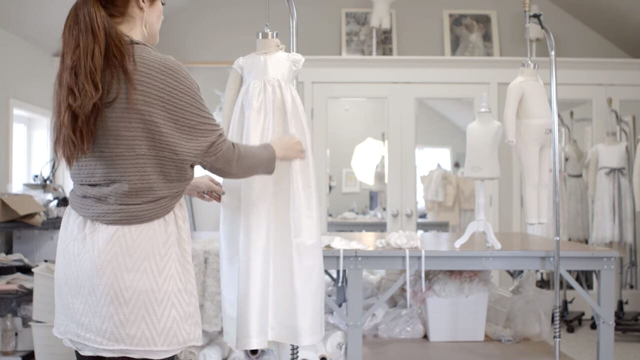 A woman with long hair is adjusting a white dress on a mannequin in a sewing room. The room is filled with sewing supplies, including tables, mannequins, and framed pictures on the wall.