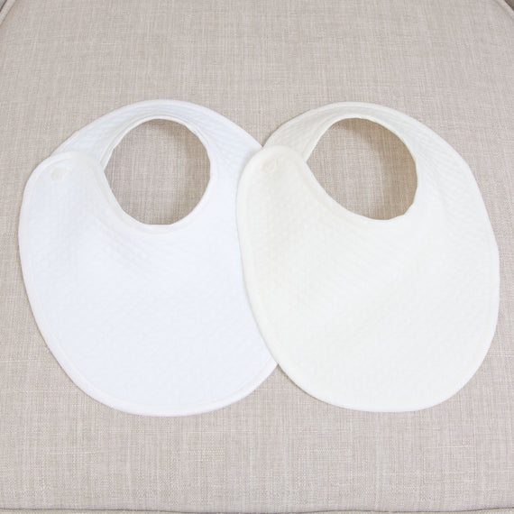 Photo color comparison of two Oliver Bibs, one in white and the other in ivory. Both are made from 100% quilted cotton and feature a button closure.