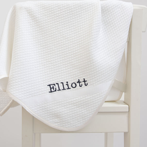 Flat lay photo of the Elliott Personalized Blanket, a soft white 100% quilted cotton, one corner is detailed with ribbon and the name "Elliott" embroidered.