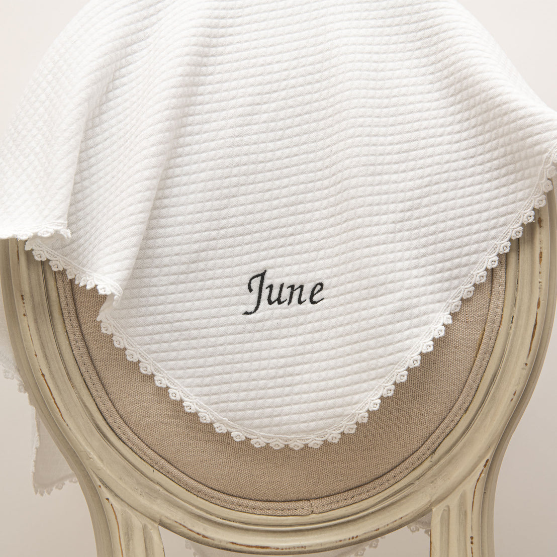 A close-up of a vintage wooden chair with a white June Personalized Blanket draped over it, featuring the word "June" embroidered in the center, surrounded by stylish lace trim.