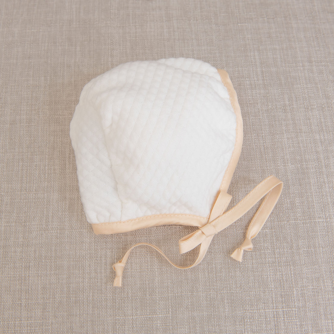 Flat lay photo of the Liam Quilted Newborn Bonnet made with soft quilted cotton in ivory and trimmed with a champagne colored silk