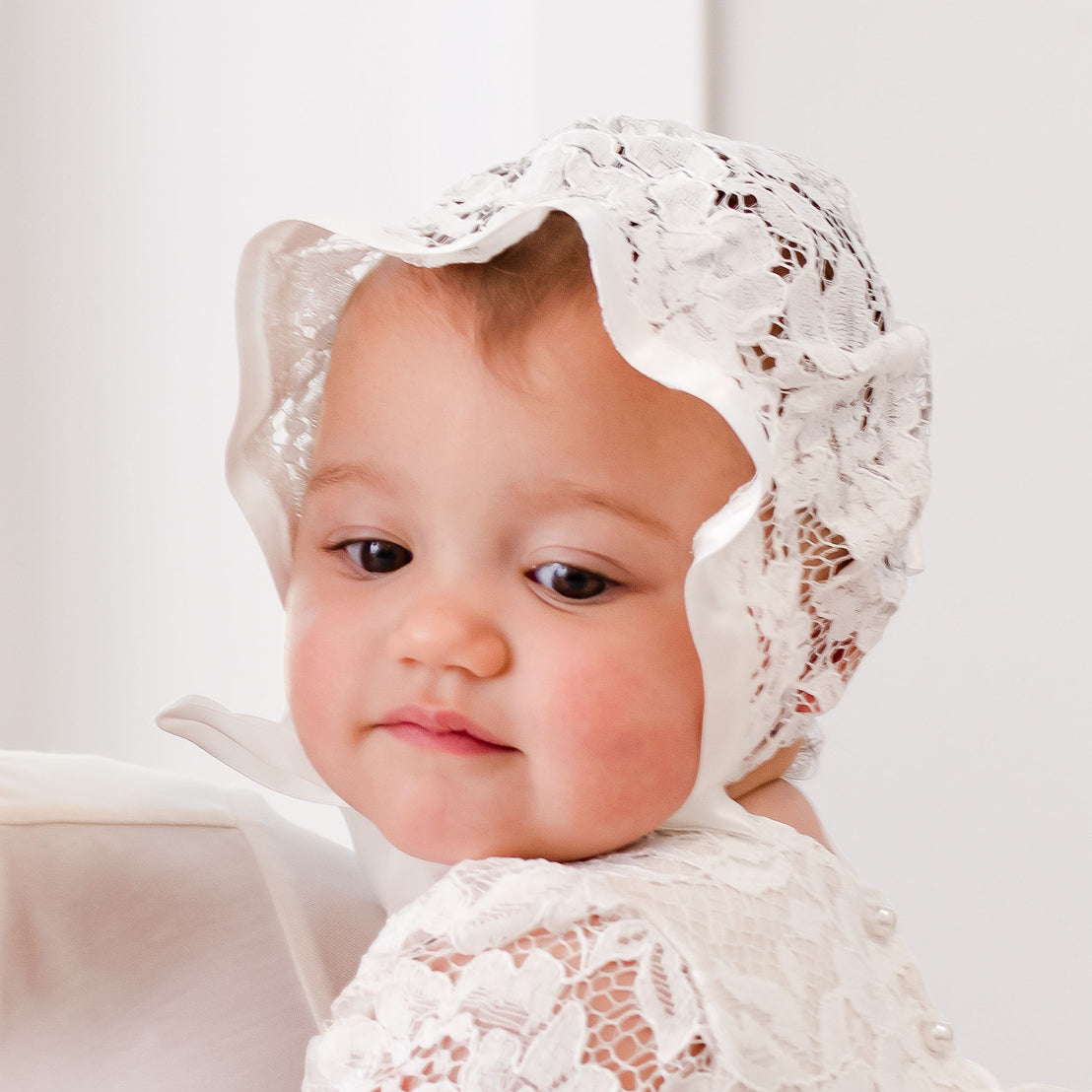 A close-up of a toddler wearing the Rose Lace Bonnet, looking slightly away from the camera with a soft, thoughtful expression. The background is plain and light-colored, focusing attention on the child.
