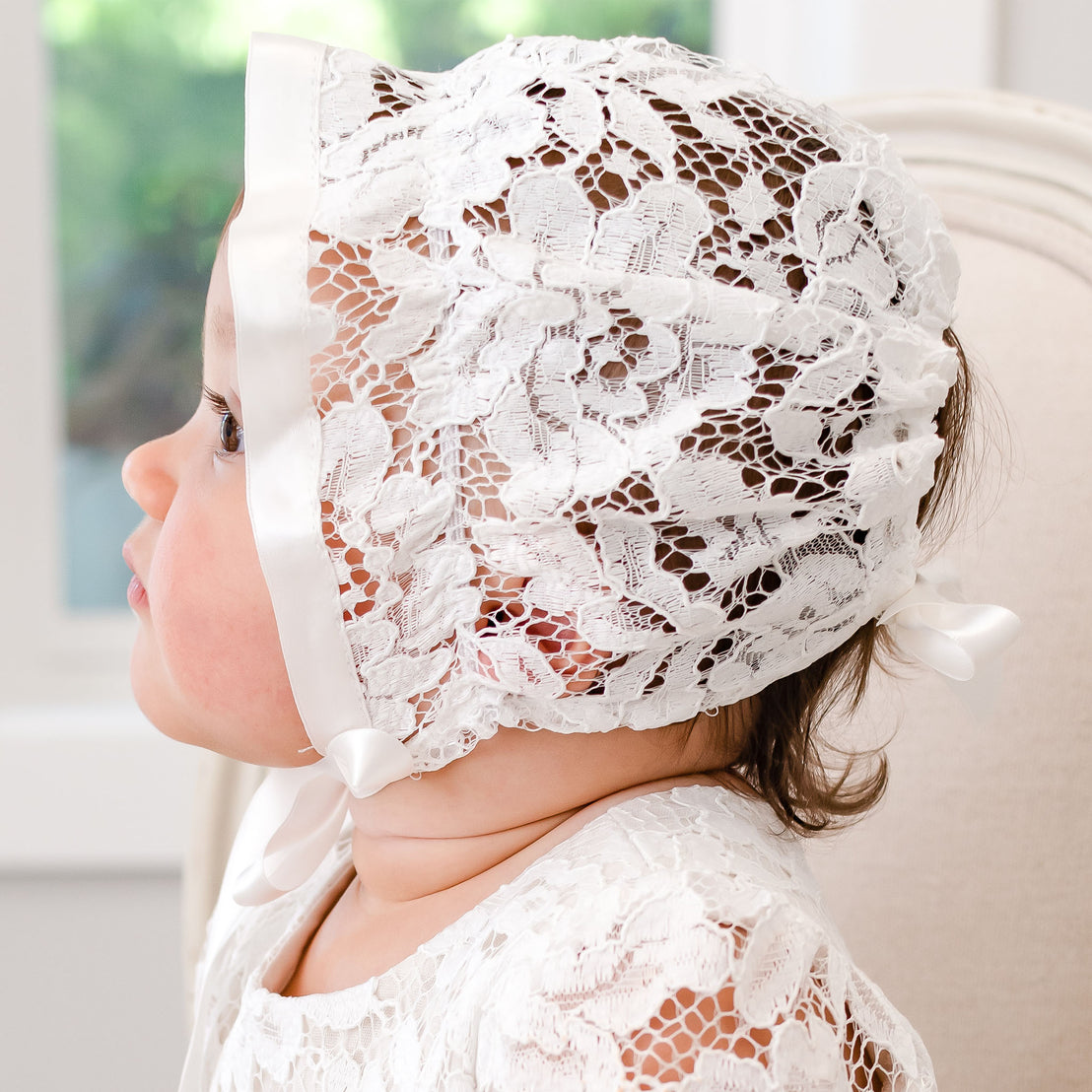 A toddler wearing the Rose Lace Bonnet and dress gazes out a window, with soft lighting illuminating her profile.