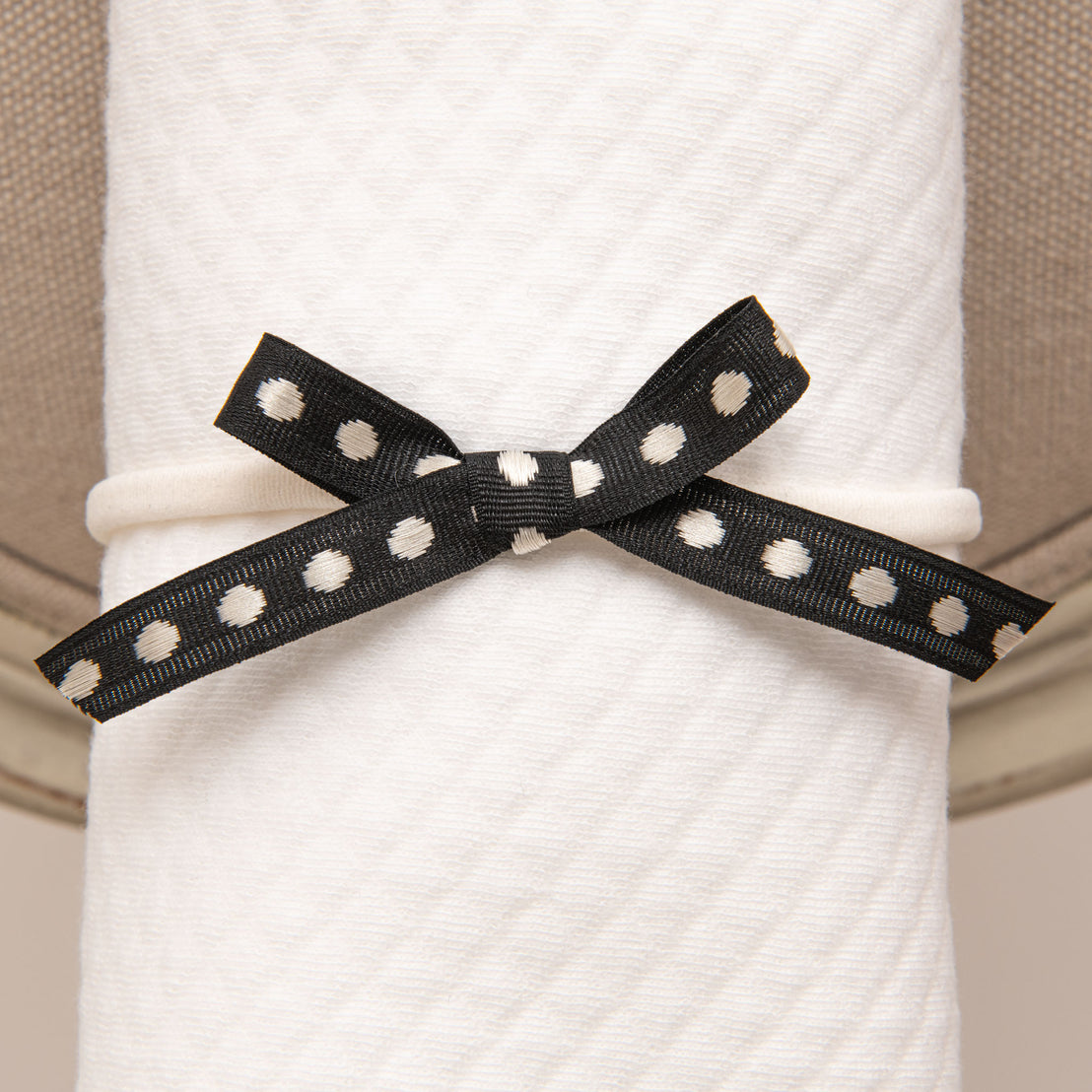 A white, textured blanket is tied with a June Bow Headband featuring small white polka dots, creating a stylish bow at the center. The background is neutral beige.