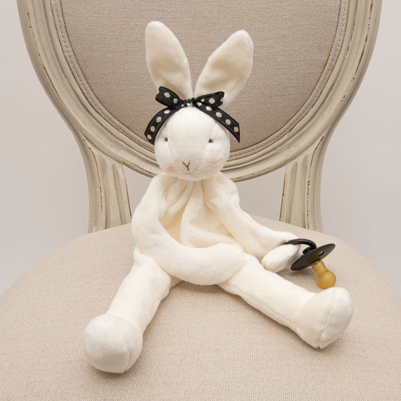 A June Silly Bunny Buddy with a stylish black polka-dotted bow on its head, sitting on a beige chair. The bunny holds a yellow and black pacifier in one hand.