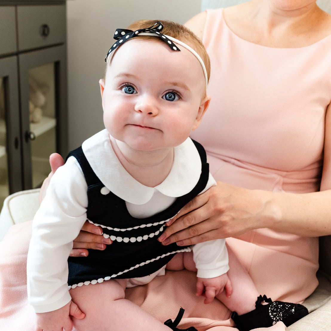 A baby with blue eyes and a stylish black and white dress sits on the lap of a woman in a pink blouse, both looking towards the camera. The baby wears black June booties and appears curious.