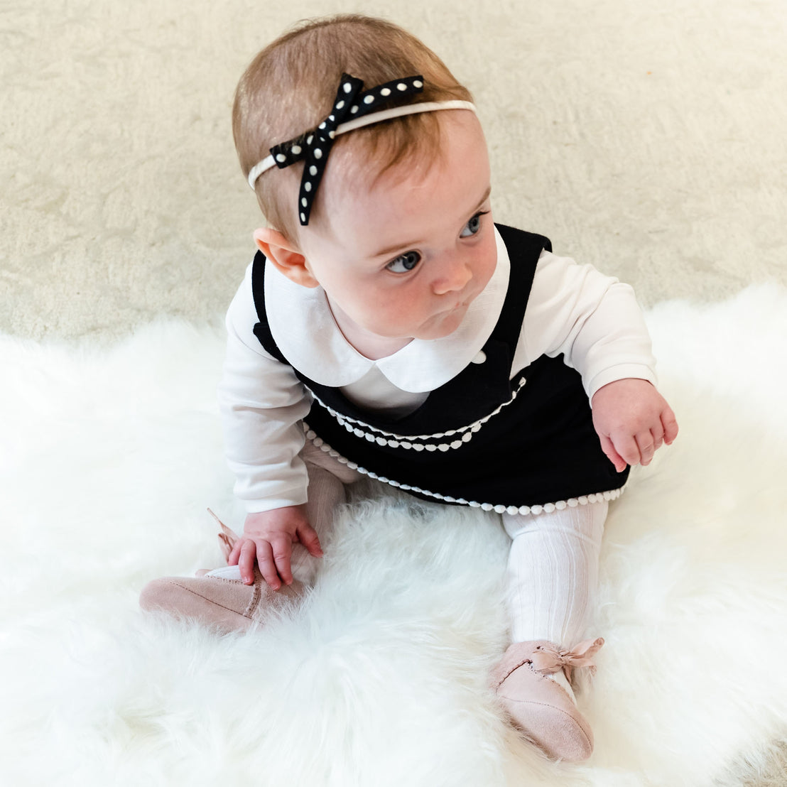 A baby girl sitting on a white fuzzy blanket, wearing a stylish black and white dress with a June Bow Headband and pink shoes, looking to her right with a curious expression.