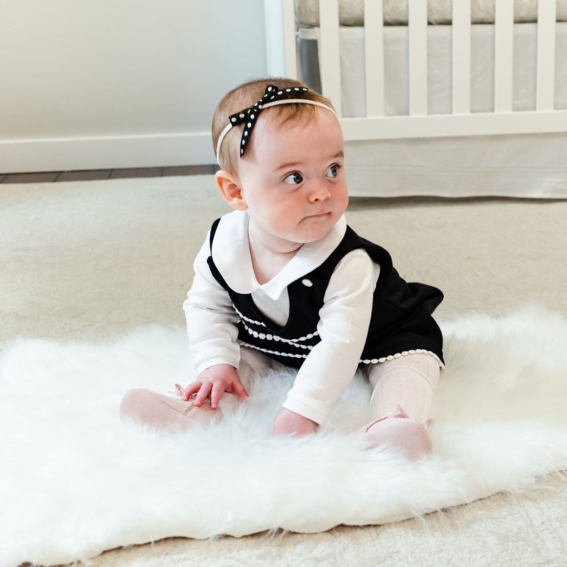 A baby girl in a stylish black and white dress and a headband sits on a soft rug, looking to the side with a focused expression, in a room with June Suede Tie Mary Janes in the background.