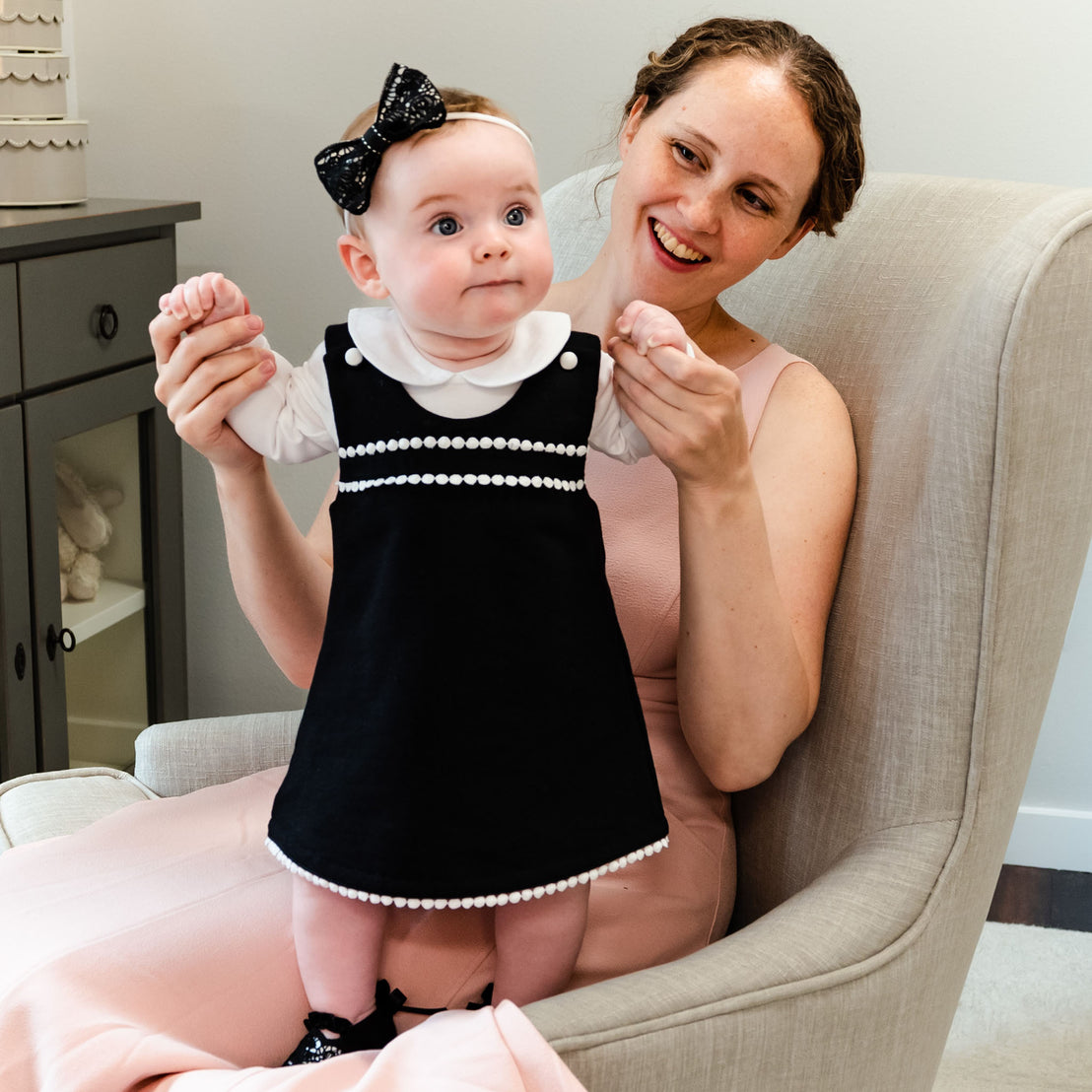 A woman in a stylish outfit smiles while sitting on a beige chair, holding a baby girl dressed in a June Jumper Dress Set with a bow headband after her christening. The baby looks surprised.