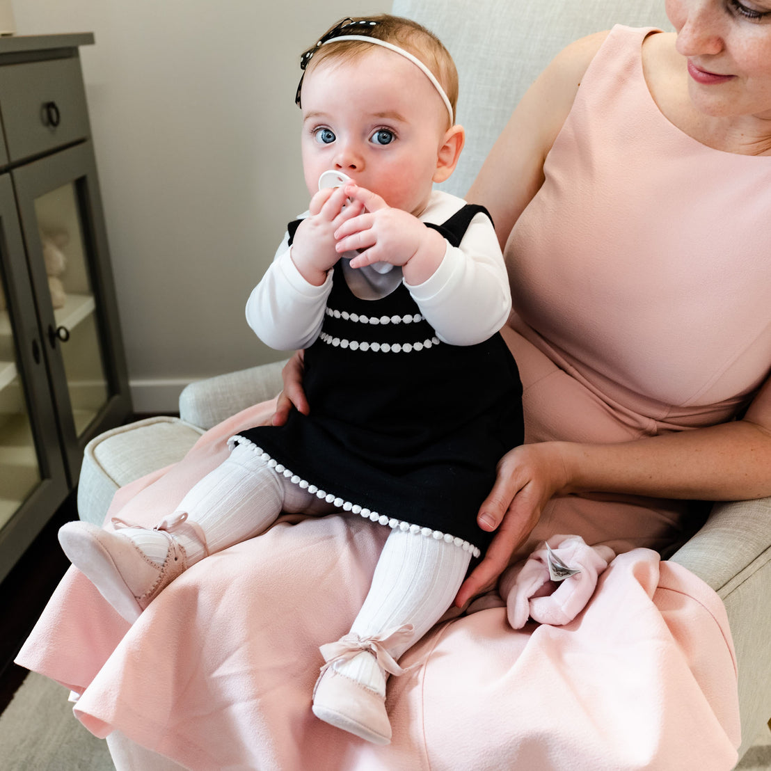 A woman in a pink dress holds a baby girl in a black and white dress after her christening; the baby is chewing on her pacifier. They are sitting in an armchair in June Suede Tie Mary Janes.