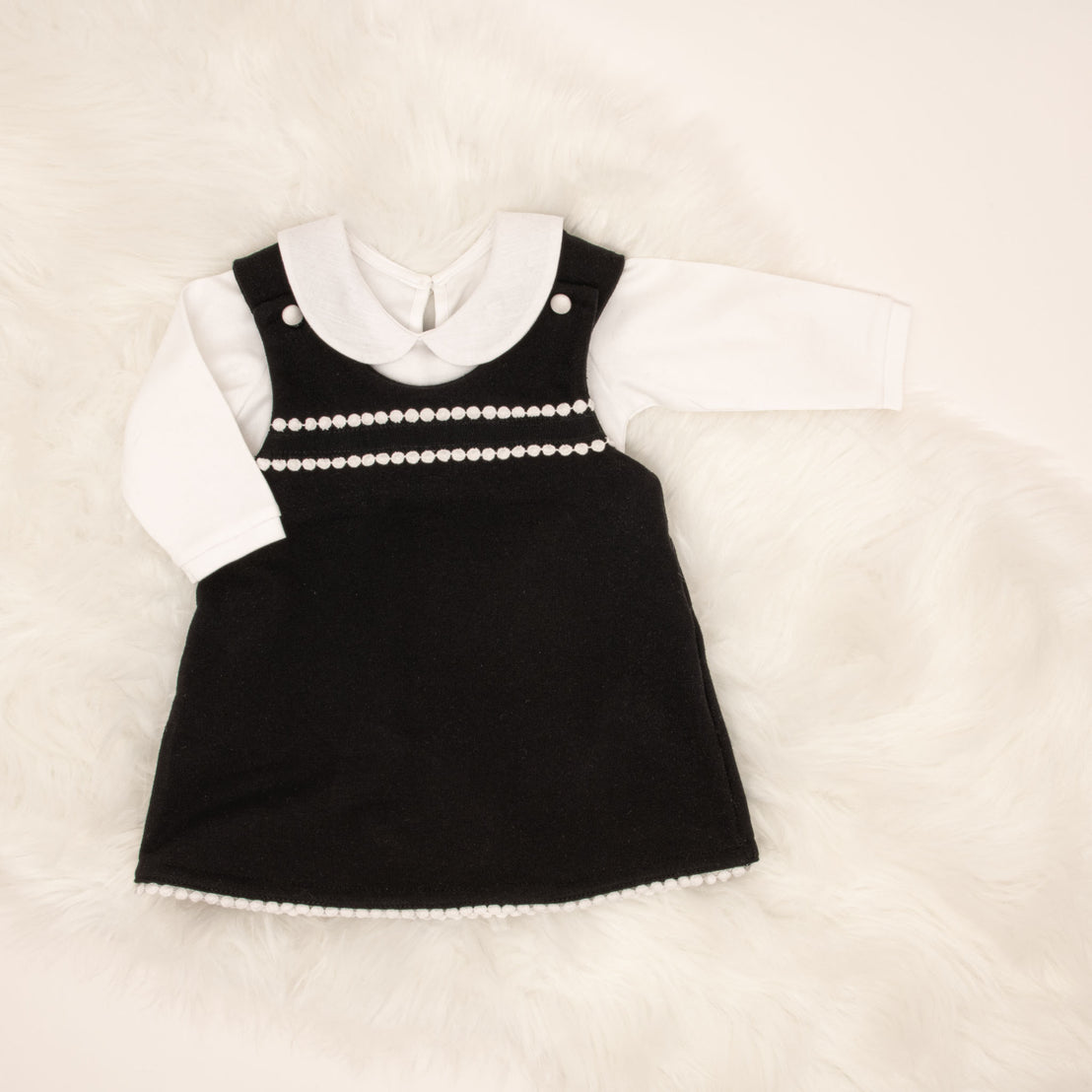 A baby's June Jumper Dress Set with white accents and pearl details for after christening, displayed on a soft, white furry surface. The dress features a layered look with a white long-sleeved shirt.