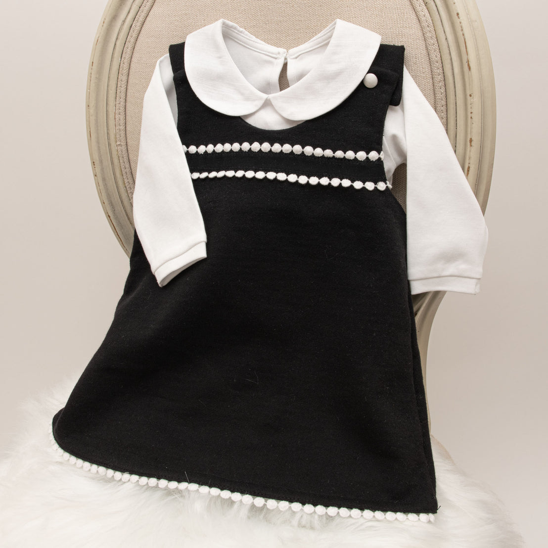 A June Jumper Dress Set with a white collar and pearl detailing on display, draped over a fuzzy, white fabric on a round chair. The dress features a simple design with decorative white pearls.