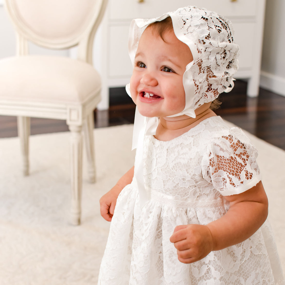 A toddler in a white lace dress and a Handmade Babydollz Rose Lace Bonnet smiles brightly while standing on a wooden floor against a backdrop of a white chair and wall.