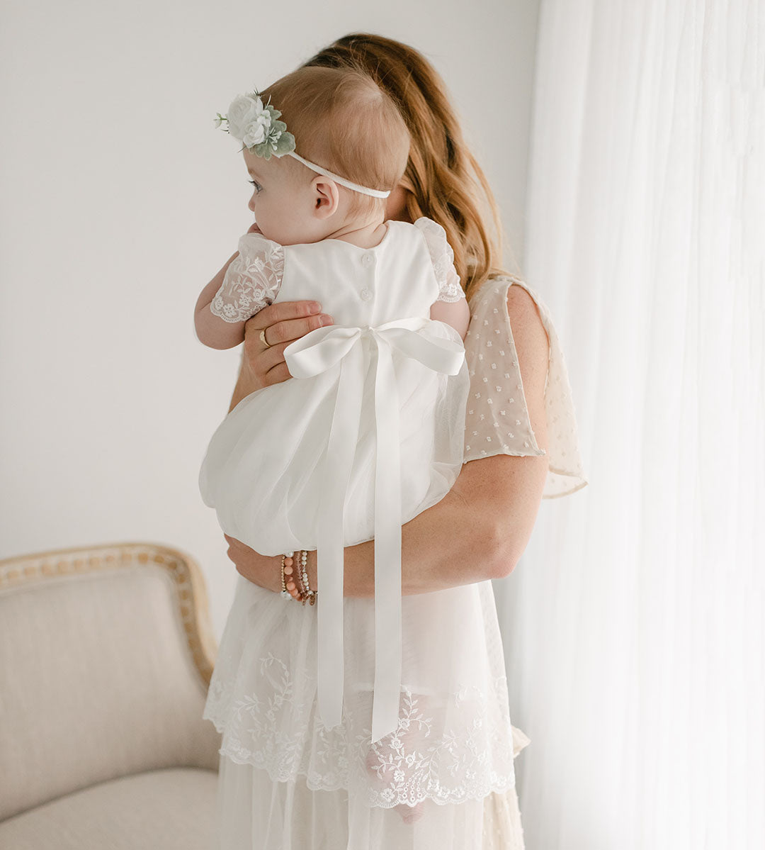 mom holding baby daughter wearing ella baptism gown with silk bow at back