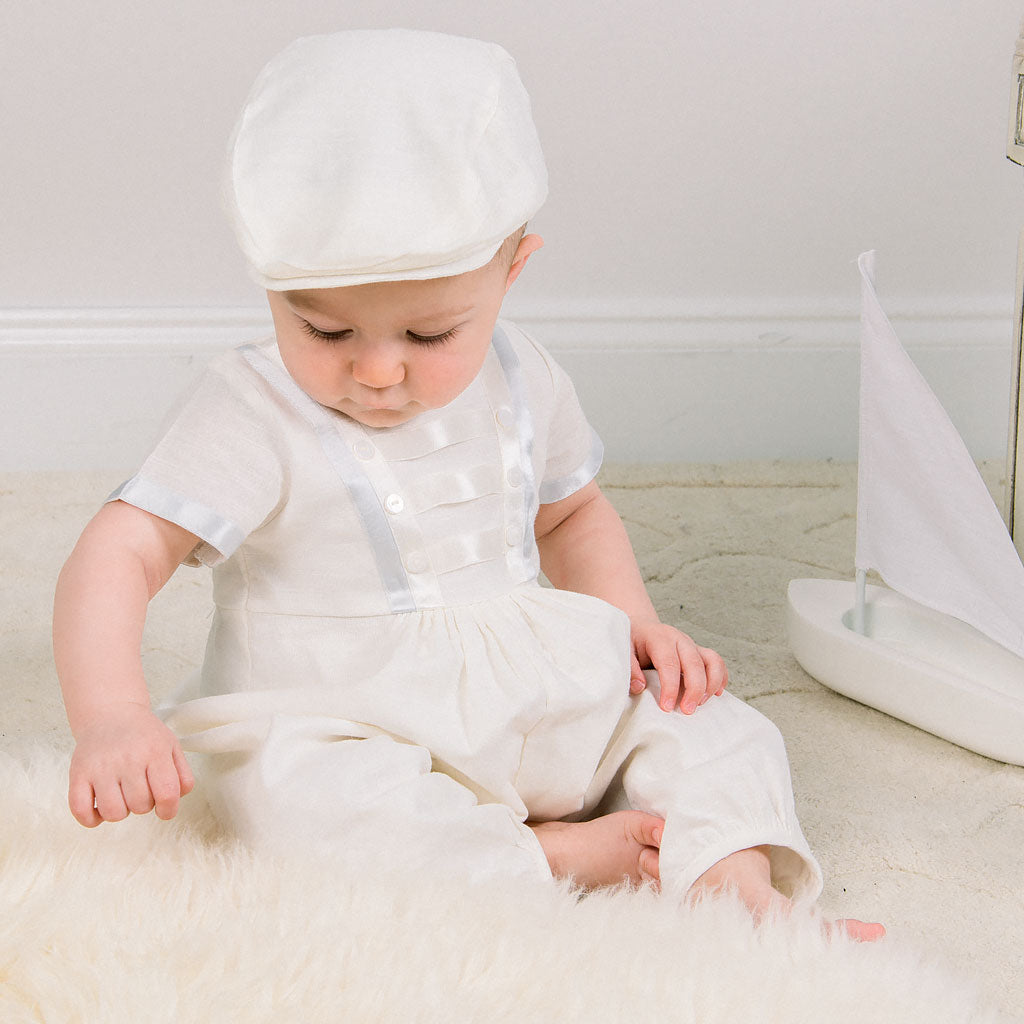 Baby boy sitting down and wearing the Owen Newsboy Cap and matching Linen Romper featuring silk ribbon in ivory and light blue across the front bodice and sleeves