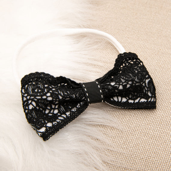 A stylish black lace June Lace Bow Headband with a delicate floral pattern, displayed on a soft, beige textured surface. The headband features thin, elastic straps and center ruching detail.