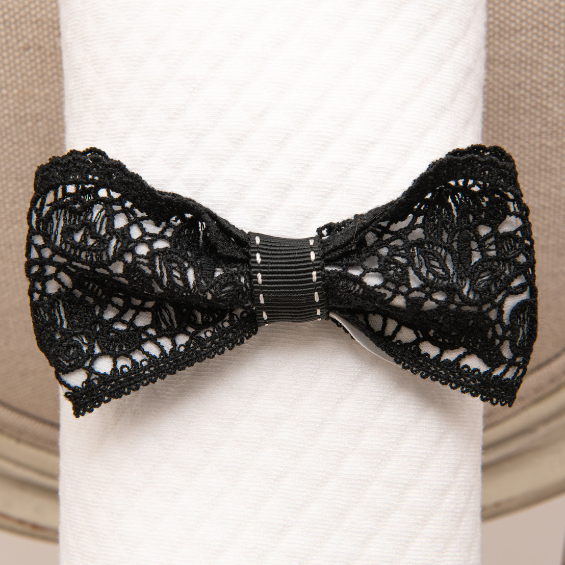 A stylish black June Lace Bow Headband clasping a white wrapped baby blanket, set against a neutral background. The bow features intricate lace patterns with scalloped edges.