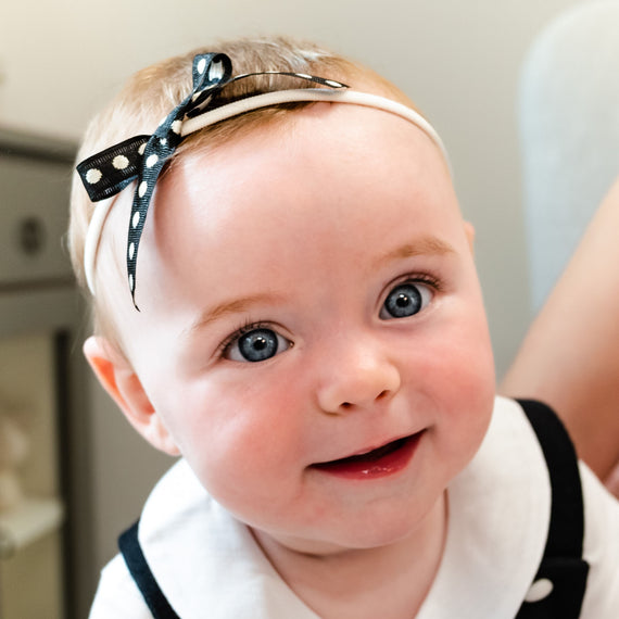 A close-up of a smiling baby with bright blue eyes, wearing a stylish June Bow Headband. The baby has rosy cheeks and is dressed in a white outfit.