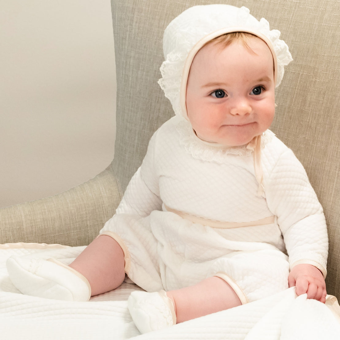 Newborn baby girl sitting on a chair and wearing the Tessa Quilt Romper and Tessa Quilt Bonnet. The romper is made from a white quilted cotton and features ivory Venice Lace along the neck and sleeves and pink champagne silk trim at the cuffs, waist and neck.