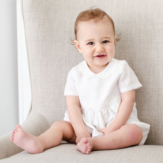 Baby boy sitting on chair and wearing the Oliver Romper, a bubble romper made from linen featuring a collar and bodice with buttons and Venice lace detail.