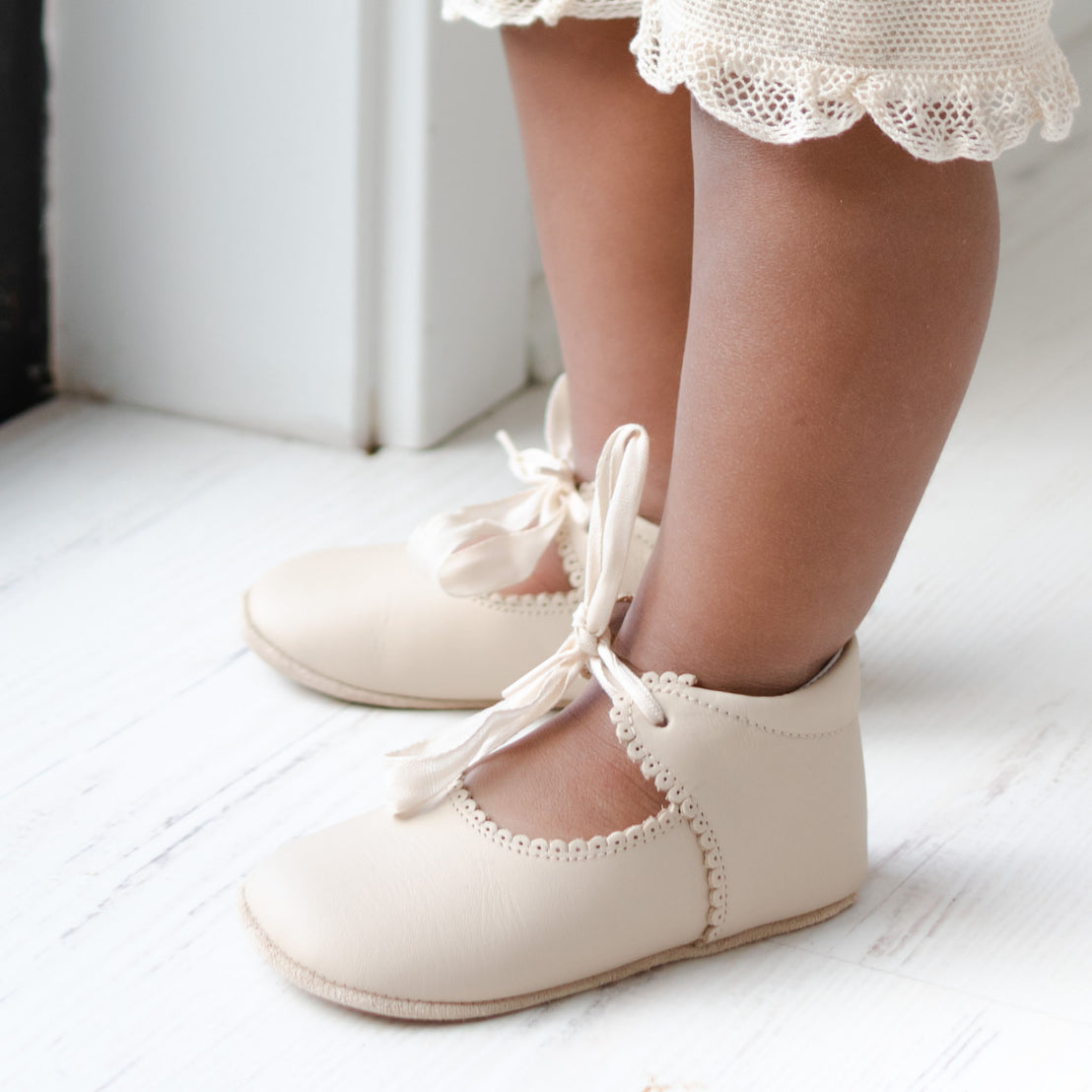Side view detail of a baby wearing the Tan Tie Mary Janes crafted with tan matte leather with beautiful scallop edge detail and complimenting cotton ribbon tie