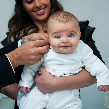 Baby being held by mom wearing a baptism romper. Baby boy is smiling at camera with a surprised look.