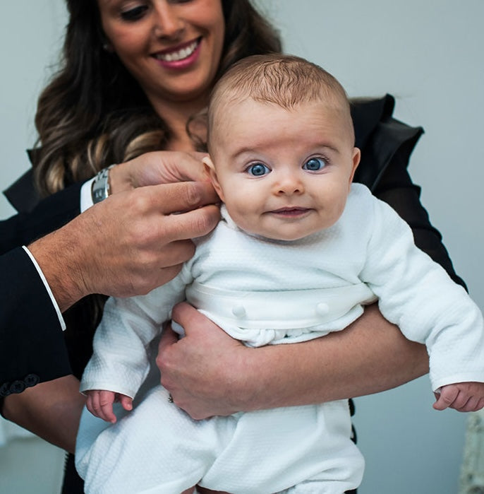 Baby being held by mom wearing a baptism romper. Baby boy is smiling at camera with a surprised look.
