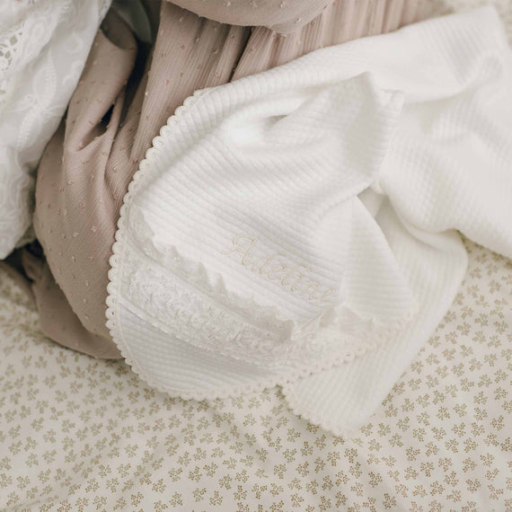The cozy scene features the Adeline Personalized Blanket, a white textured baby blanket with an embroidered name, draped over a soft, beige quilted cotton quilt that boasts a subtle geometric pattern. The neutral tones enhance the inviting atmosphere.