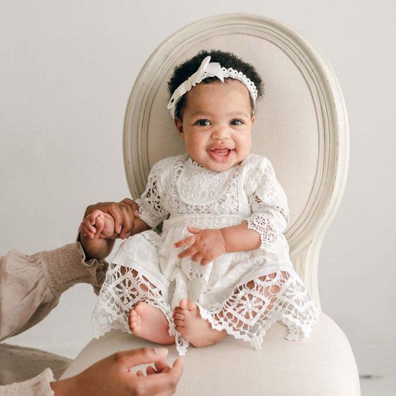 A baby sits on a white chair wearing the Adeline Lace Christening Dress & Bloomers and a headband with a bow. The baby is smiling and holding an adult's hands that are reaching in from the bottom left corner of the image, showcasing the delicate beauty of her christening dress.