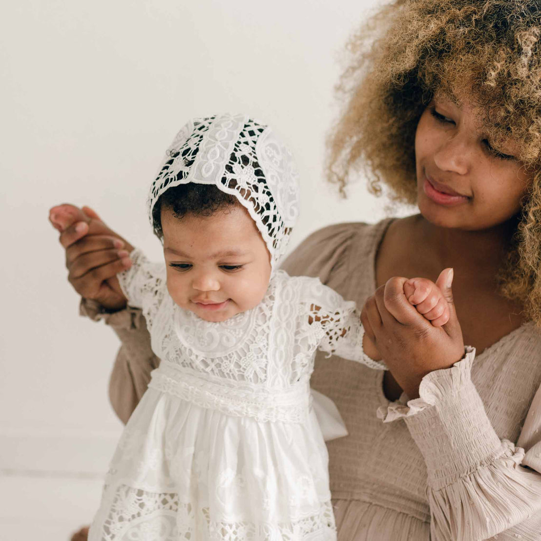 A woman with curly hair holds both hands of a baby dressed in the Adeline Lace Christening Gown & Bonnet. The baby, adorned in this white lace outfit and bonnet, stands on the woman's lap as they both look downwards. The background is light and neutral.