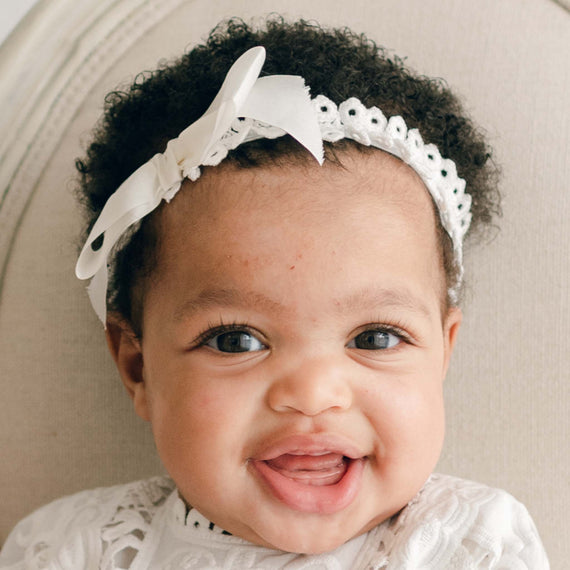 A baby with curly hair, wearing the Adeline Headband adorned with a bow, smiles while looking at the camera. The infant is dressed in a white lace Christening outfit and is seated on a light-colored chair.