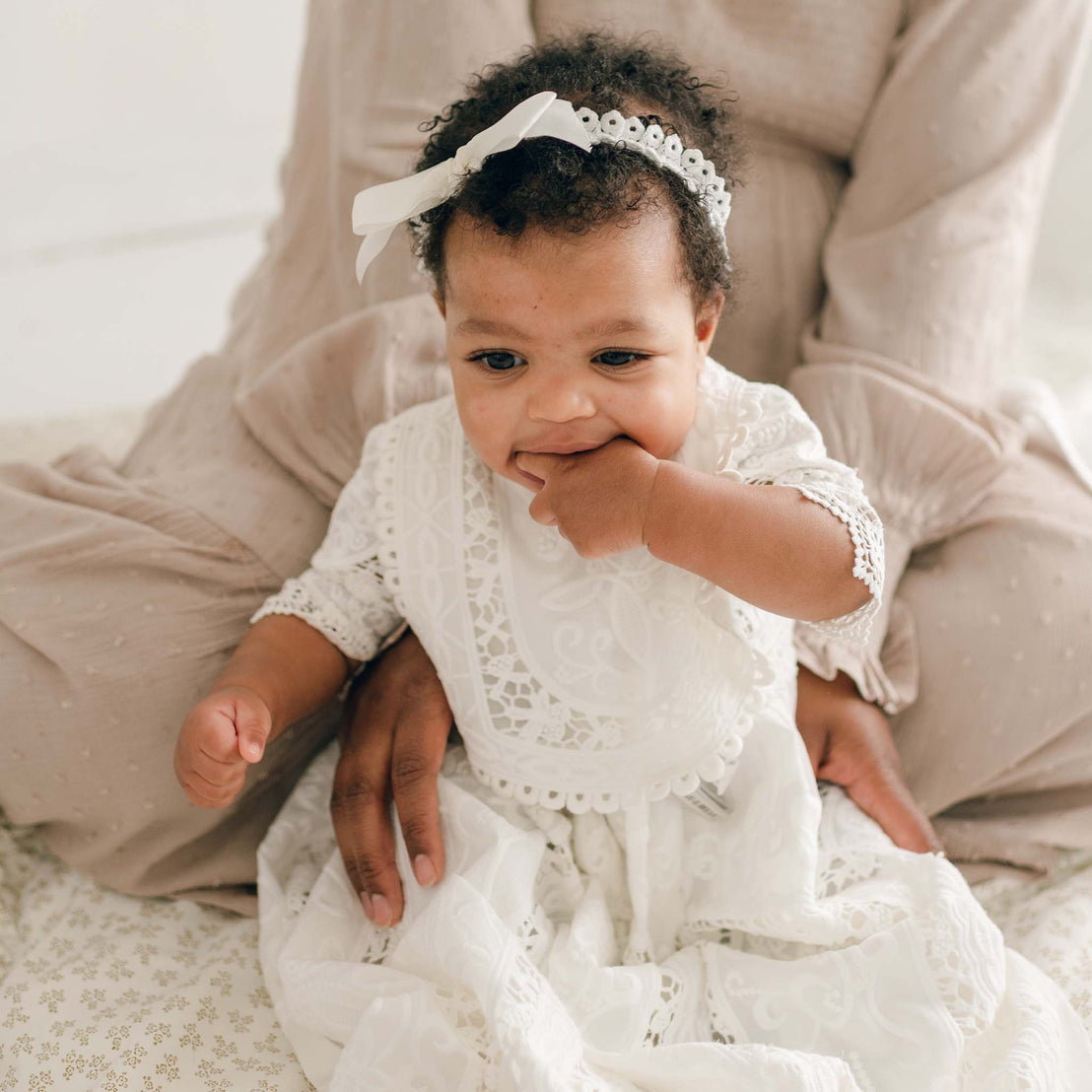 An infant in a white Adeline Bib christening dress, adorned with delicate lace details, sporting a matching white headband, is seated on a woman's lap. The baby has one hand in their mouth while the other rests by their side. The woman, dressed in a light beige outfit and mostly out of frame, gently holds the baby steady as the intricate cotton lace softly glistens.