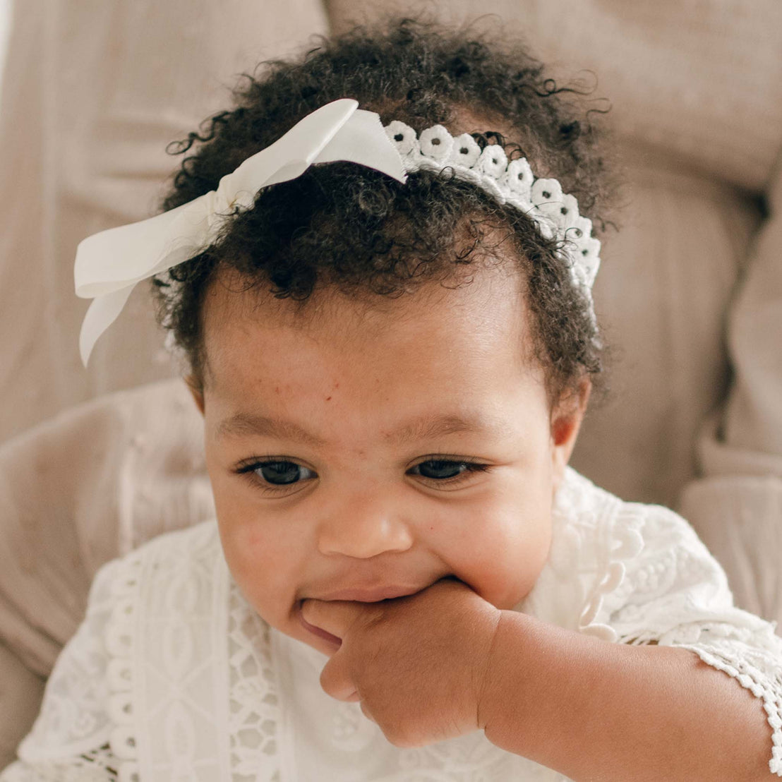 A baby with curly hair, wearing a white handmade Adeline Headband with a bow and a lacy christening outfit, has one hand in their mouth. The baby is sitting on a cushioned surface in a neutral-colored room.