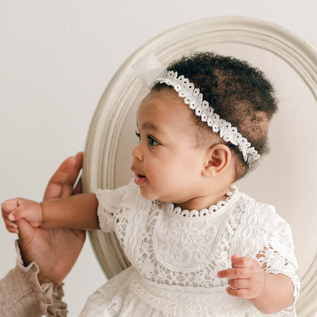 The baby, adorned in a white lace christening outfit and an Adeline Headband complete with a handmade bow, sits gracefully on a round-back chair. Looking to the side with one hand reaching out, the scene captures an adult hand gently supporting the baby from the left side of the image. This tender moment feels like a cherished gift, immortalized through love and care.