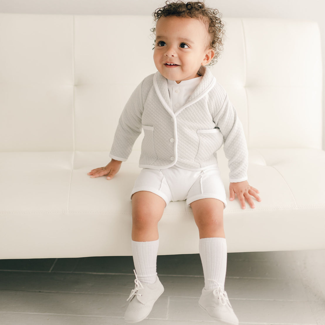 Baby boy sitting on a white couch and wearing the grey Asher 3-Piece Suit, including a folded collar jacket, white pants and a onesie with buttons.