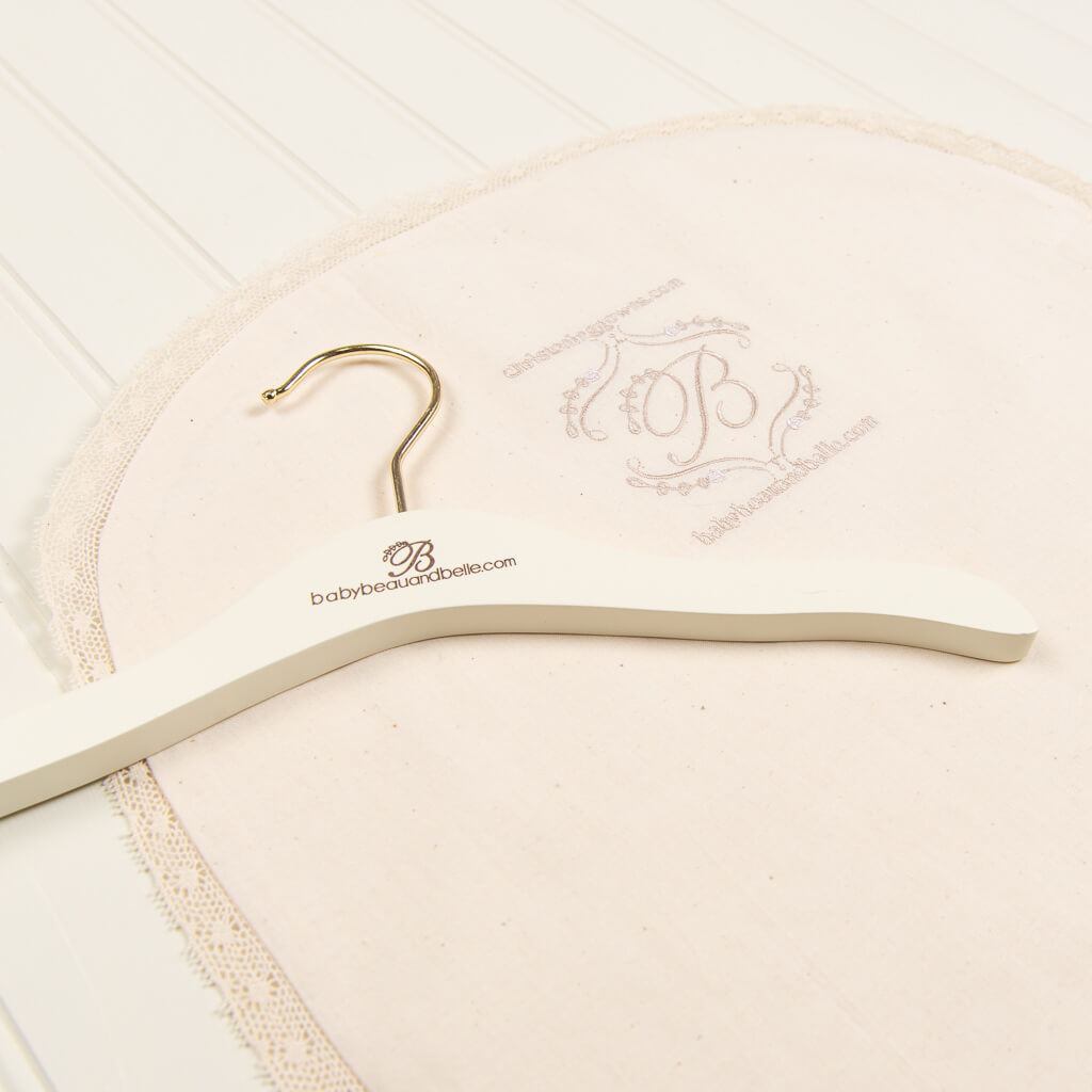 An embroidered cloth with lace trim is displayed on a white surface. A white wooden hanger marked with "babybeauandbelle.com" and a logo featuring a cursive "B" rests on top of the cloth.