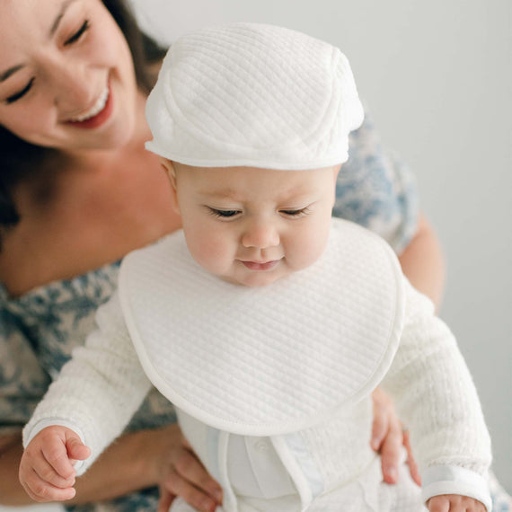 Baby boy with his mother. He is wearing the Owen Bib (and matching Owen Cotton Newsboy Cap) made out of ivory quilted cotton and an ivory linen trim.