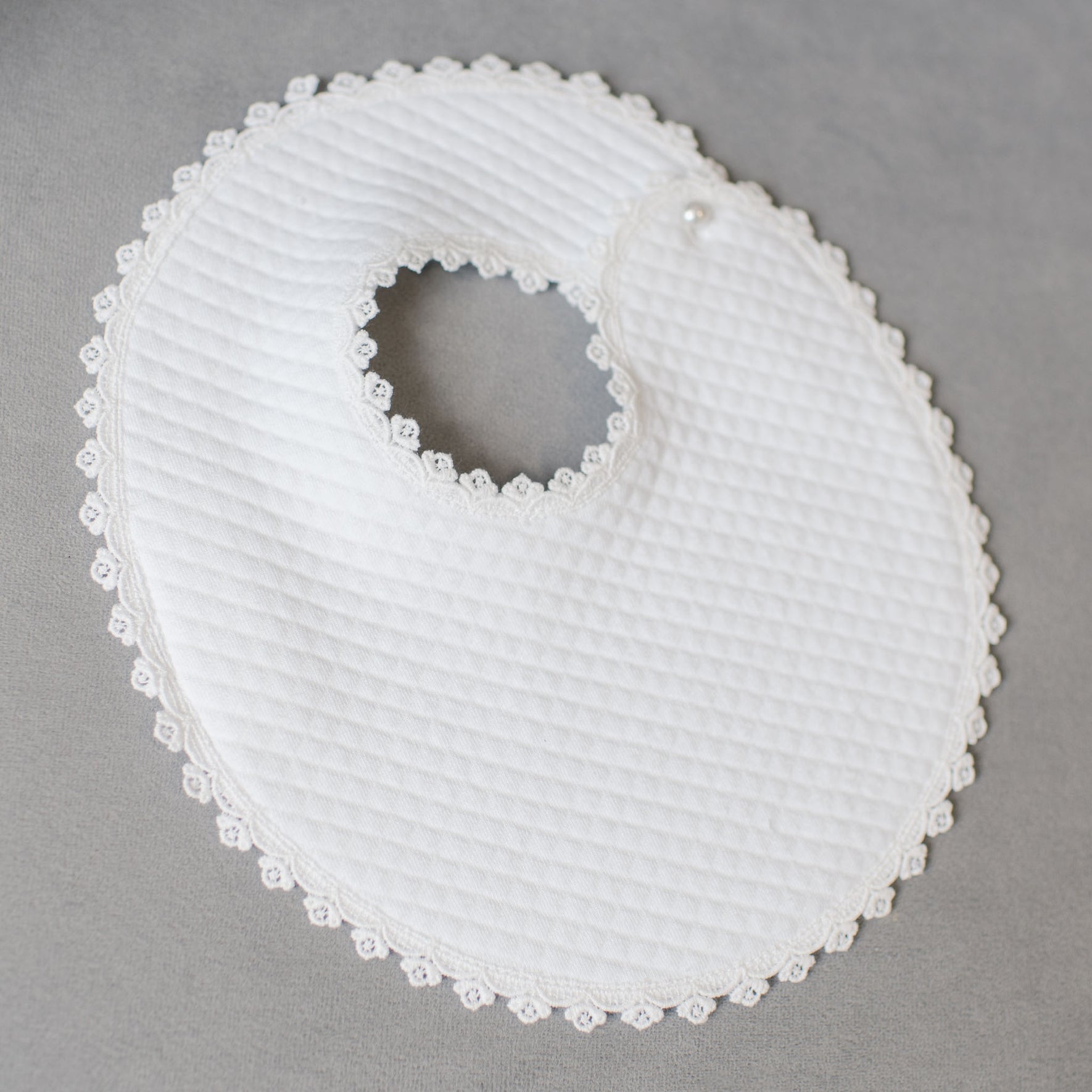 The Aria Bib is displayed on a gray surface, showcasing its round shape and white textured fabric complemented by a trim of Venice lace. The bib exudes the elegance of a christening gown, with a circular neck opening and a small decorative button near the closure, capturing the essence of designer baby attire.